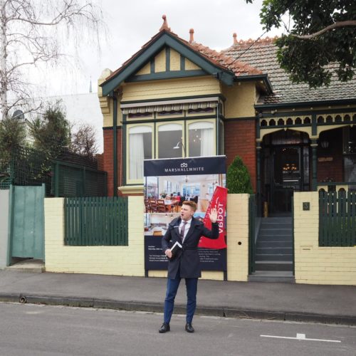 Auction snapshot - Auctioneer standing in front of yellow and red brick house