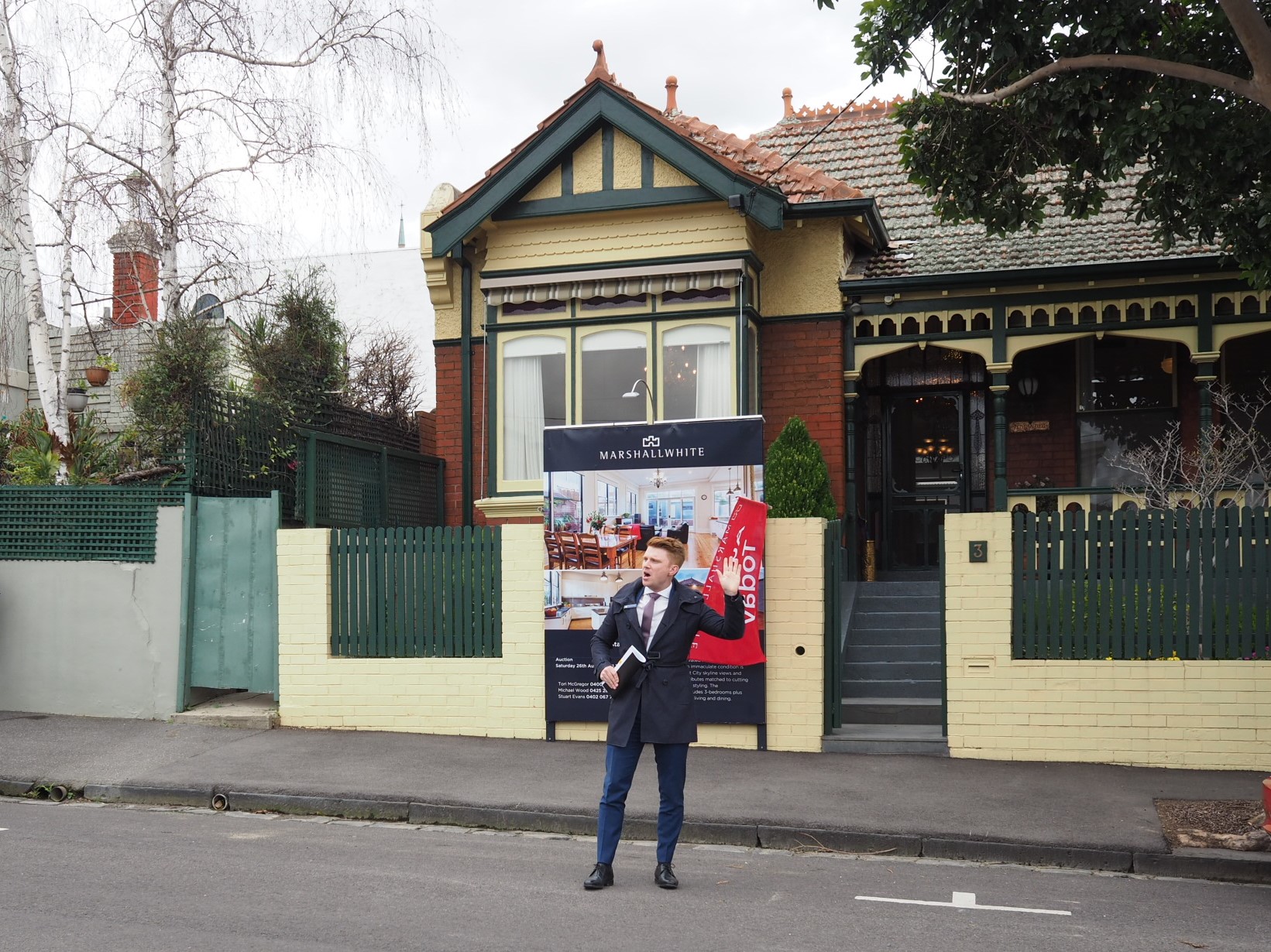 Auction snapshot - Auctioneer standing in front of yellow and red brick house